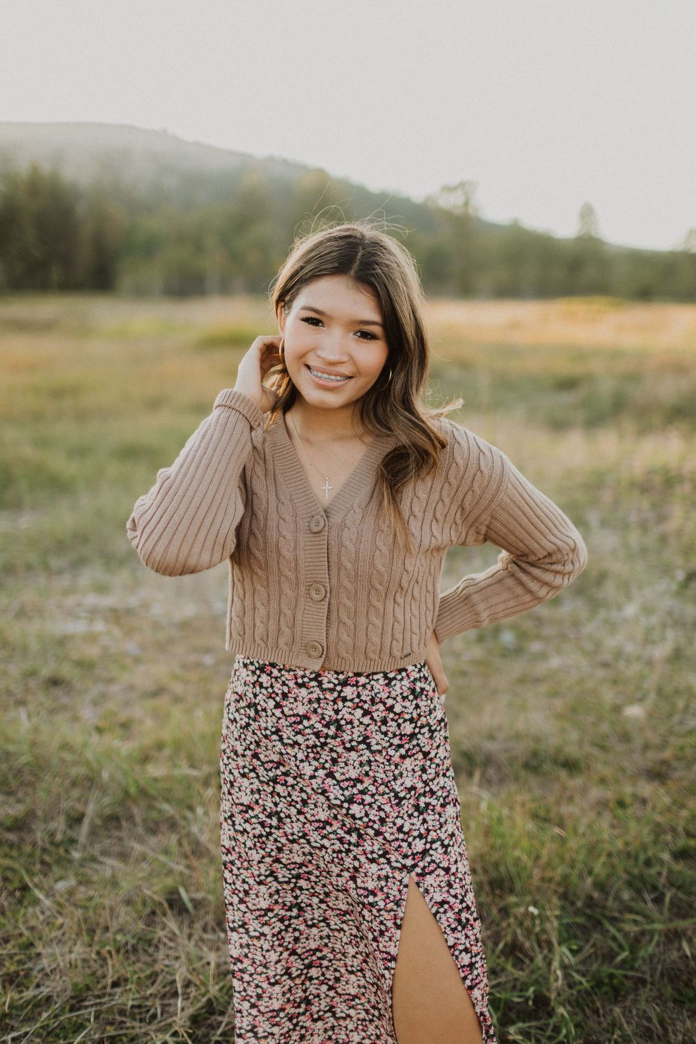 Person in tan sweater and floral skirt poses in sunny outdoor field setting.