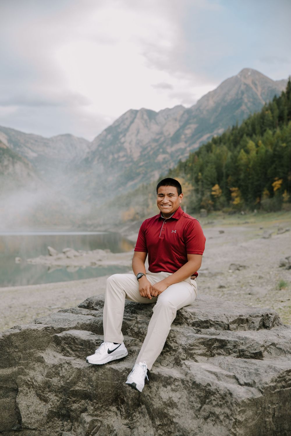 Person in red shirt sits on rocky outcrop with misty mountain landscape behind them.