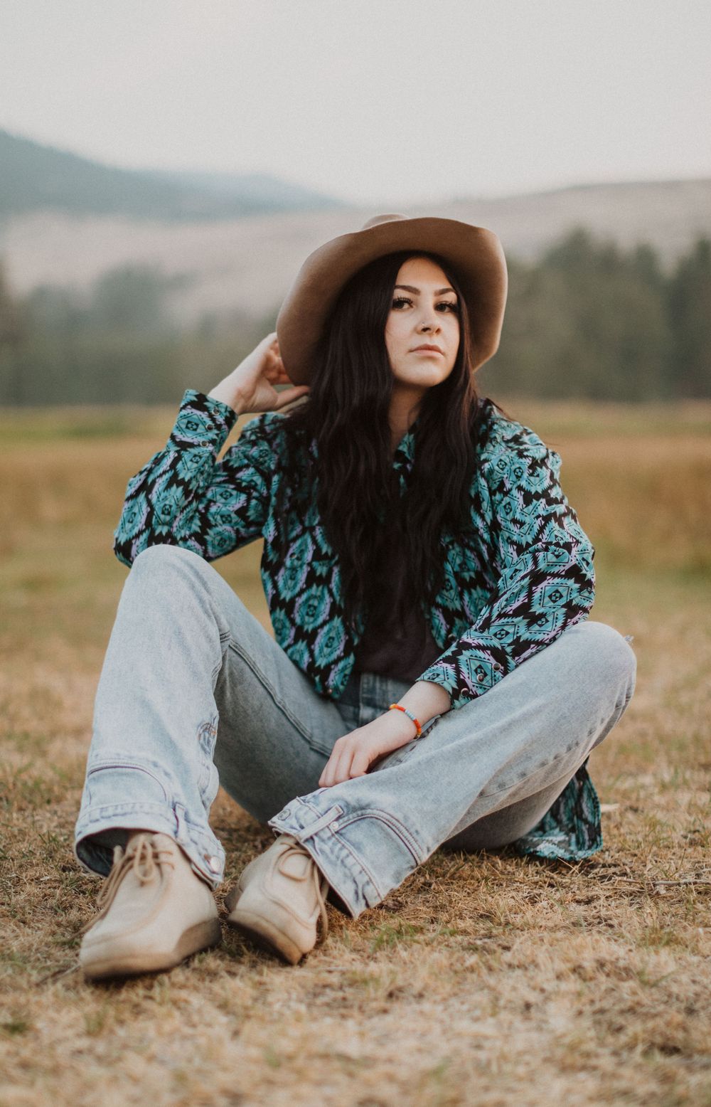 Person in floral top and jeans sits casually in a field wearing a wide-brimmed hat.