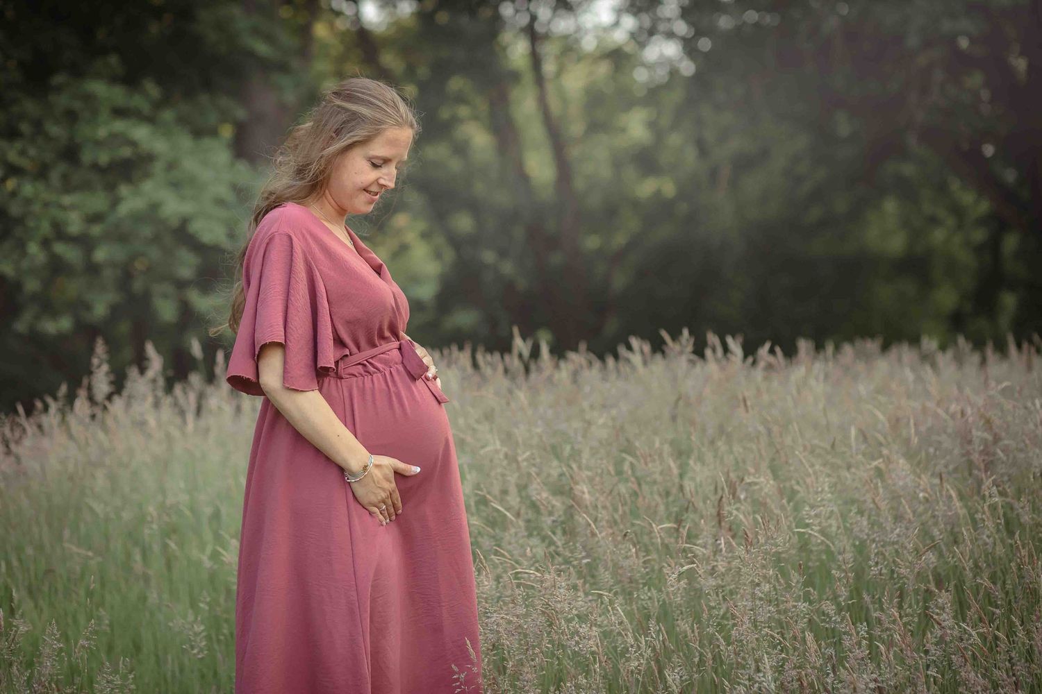 Zwangere vrouw in een roze jurk staat in een grasveld, handen liefdevol op haar buik.