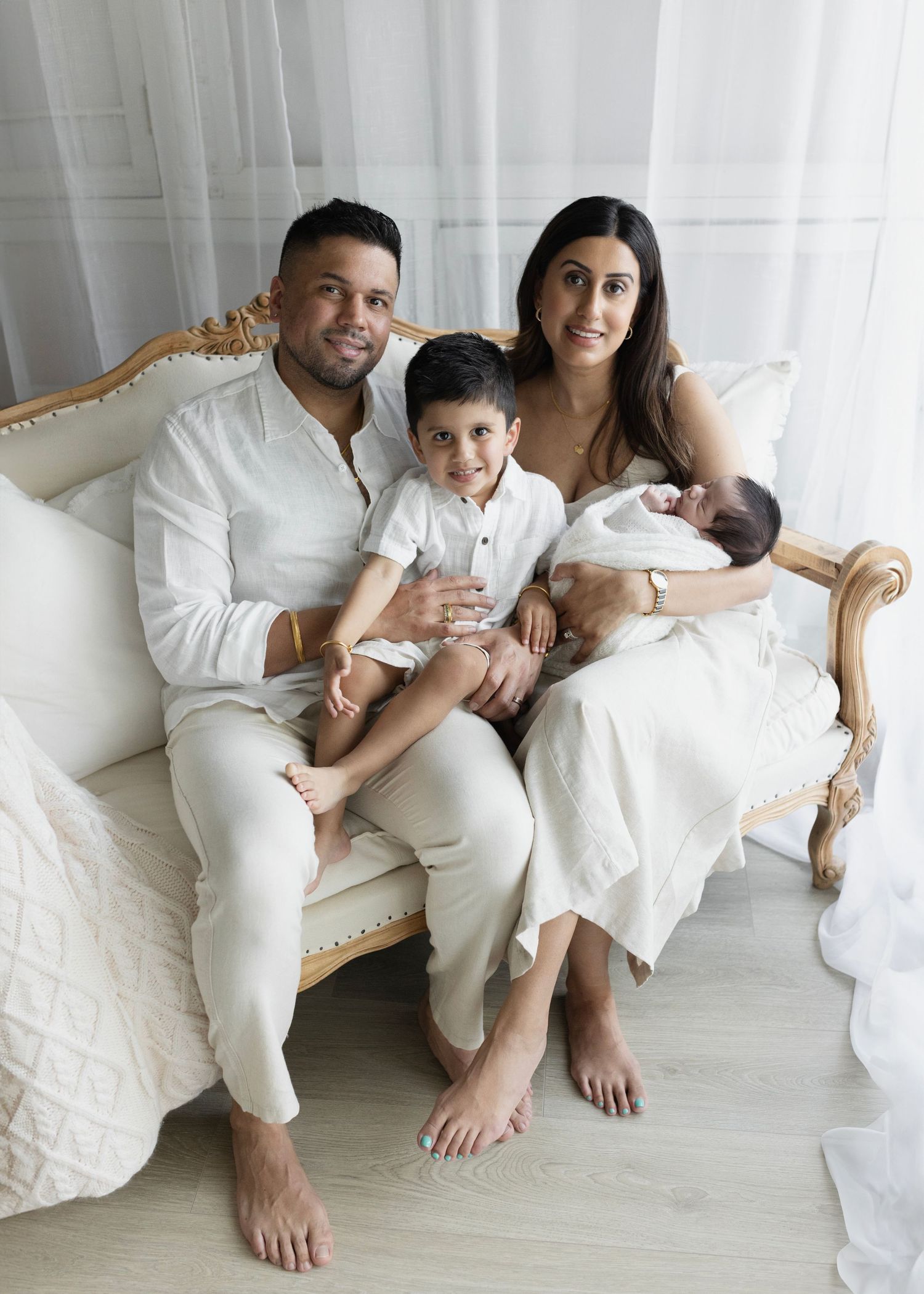 mom and dad sit on a white sofa in an all white setup with their newborn son and older son. Elizabeth Lang Photography