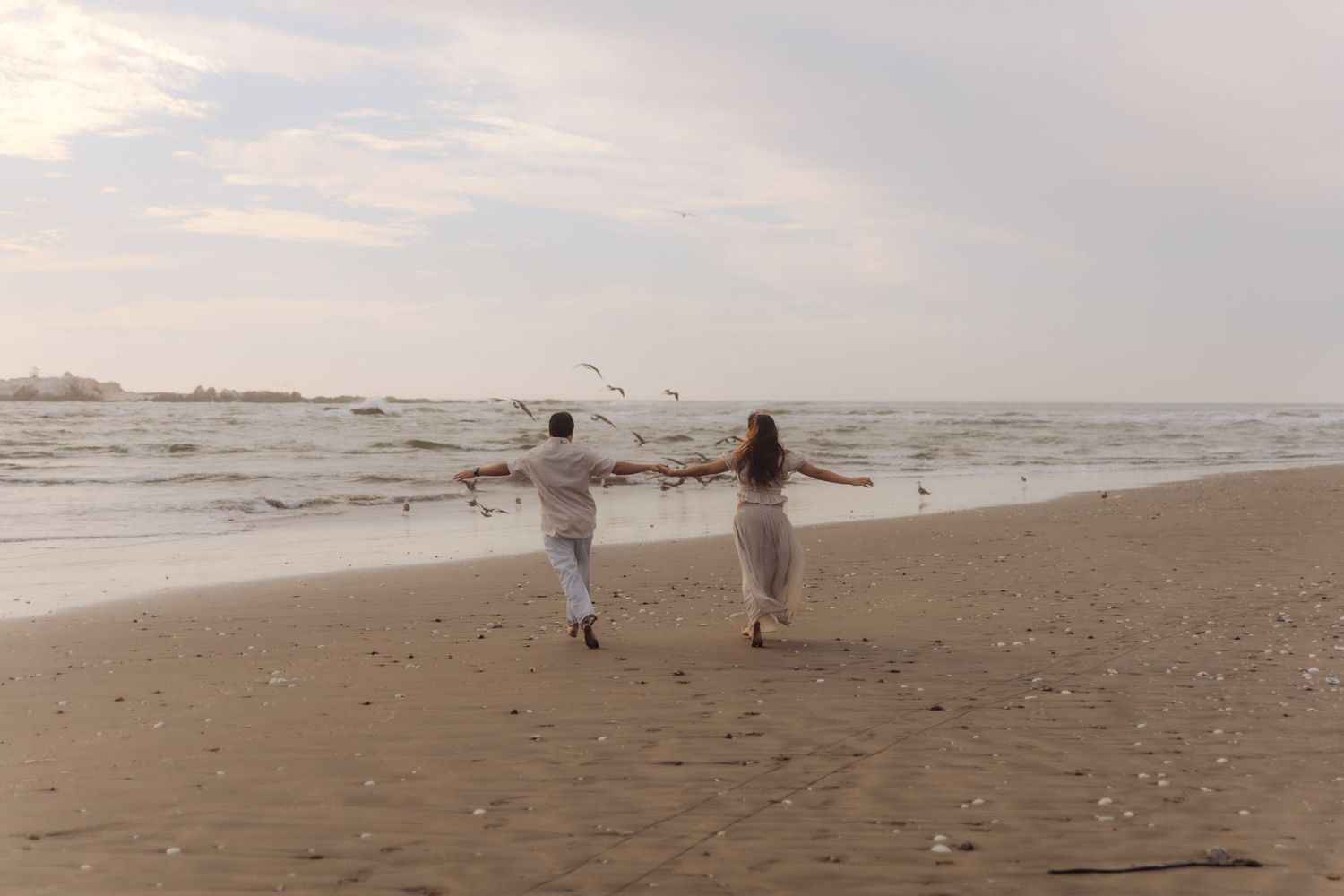 Couple holding hands running along a sandy beach with ocean waves and seagulls in the background.