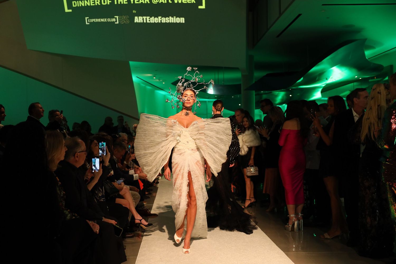 Fashion model walks runway in dramatic beige cape under green lighting at fashion show event.