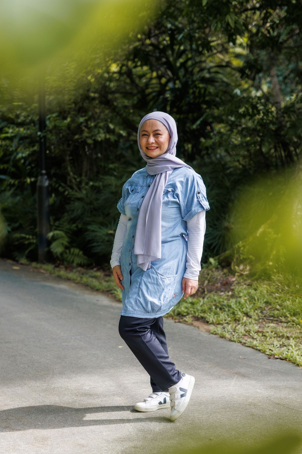 A person wearing a light blue hijab and matching tunic walks along a paved path surrounded by greenery.