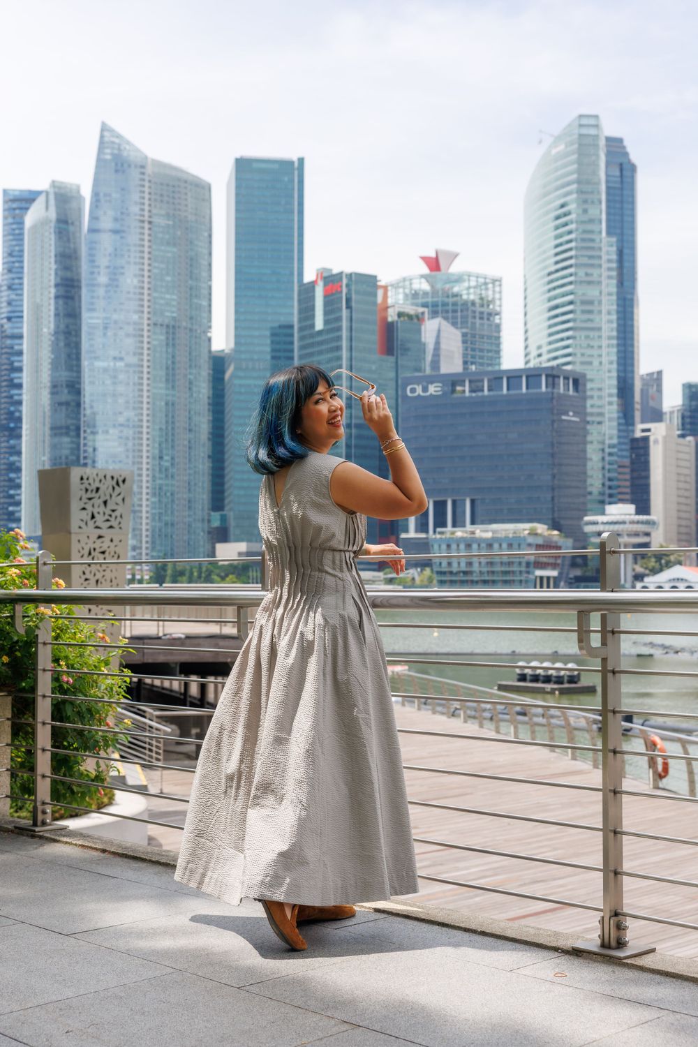 Woman in gray dress poses against Singapore's modern cityscape with skyscrapers.
