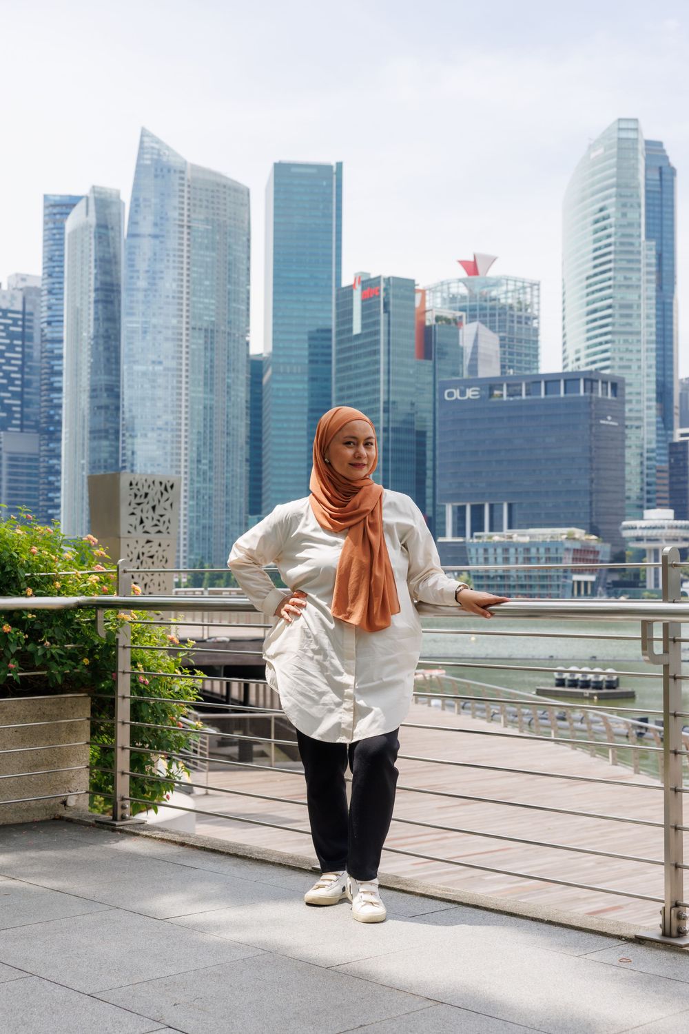 Woman in hijab poses stylishly against Singapore skyline backdrop with modern buildings.