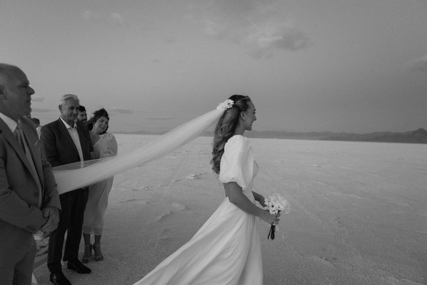 A black and white photo shows a bride in a flowing dress and veil on a beach as wind catches the fabric dramatically.