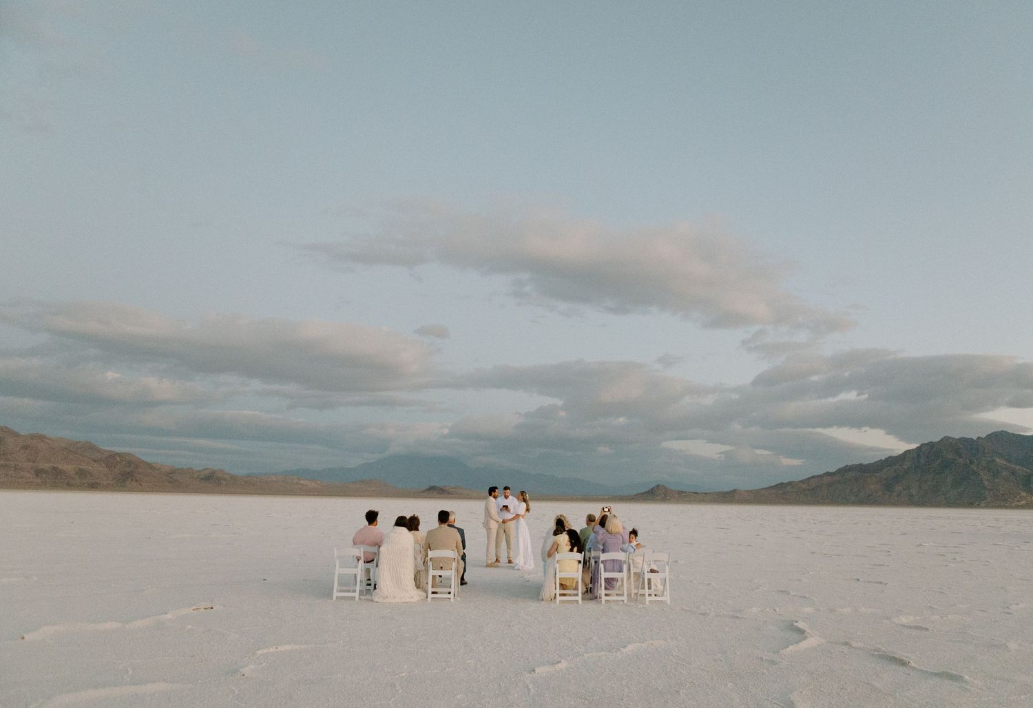 Family sit to watch a bride and groom marry on a stark salt flat landscape under cloudy skies at dawn.