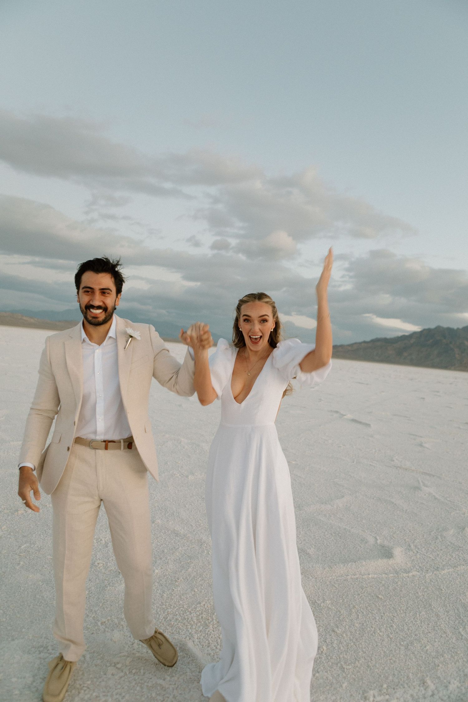 Couple in white wedding attire celebrating joyfully on white sandy beach with cloudy sky background.