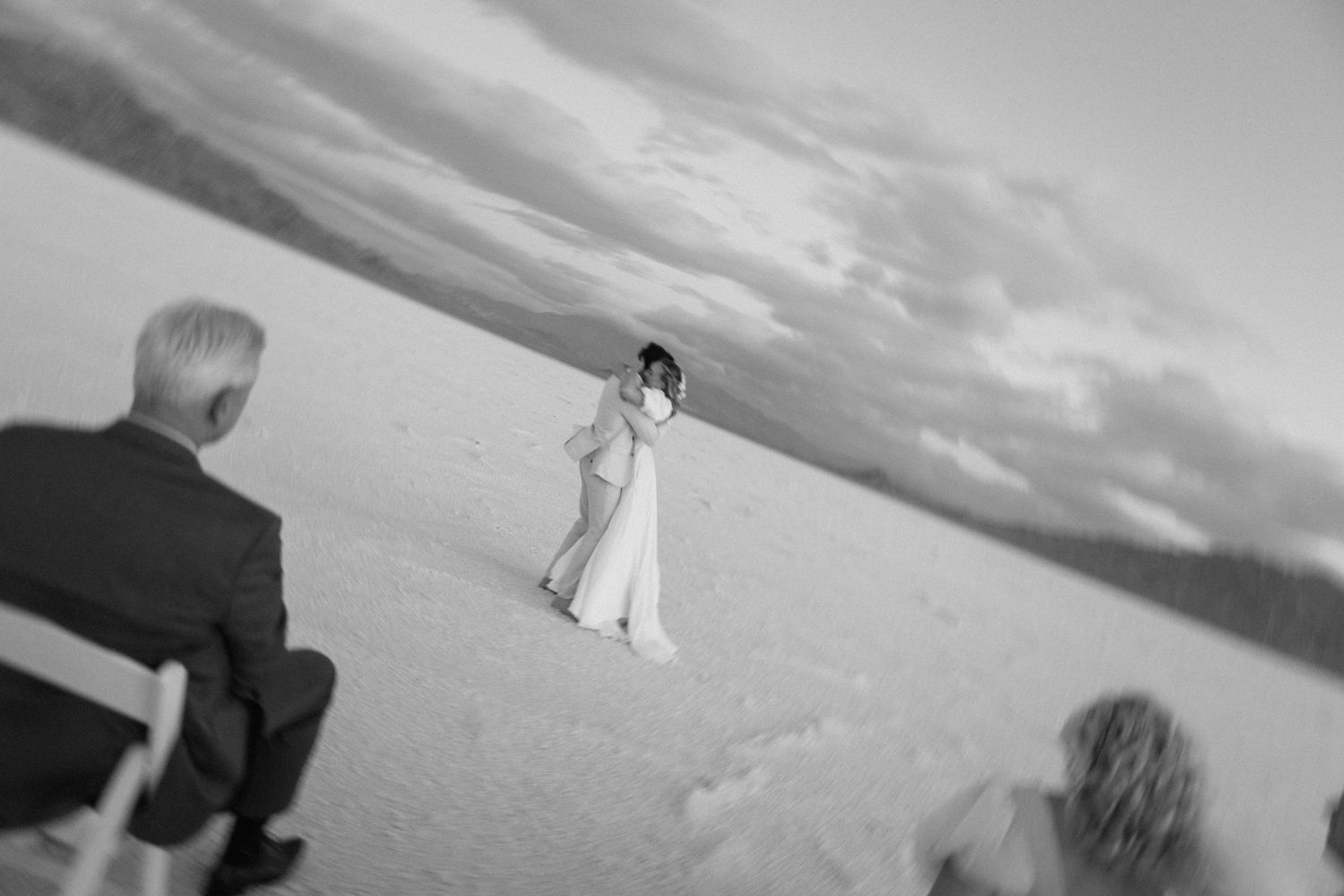 A black and white wedding photograph showing a person in a white dress walking across a salt flat with dramatic clouds above.