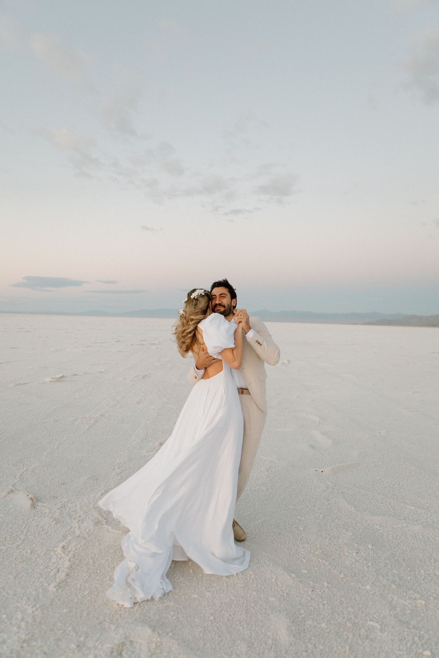 A couple in light colored wedding attire dancing on a salt flat at sunset.