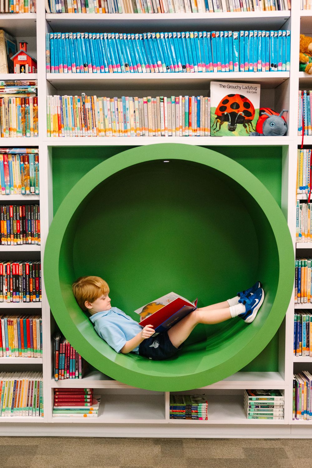 Child reading book while relaxing in green circular reading nook surrounded by library shelves.