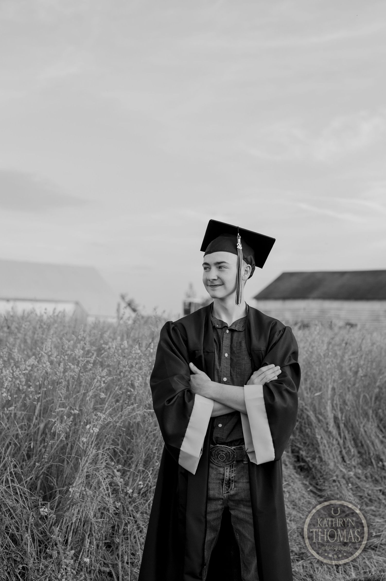 A black and white portrait of a graduate in academic regalia standing in a rural field.