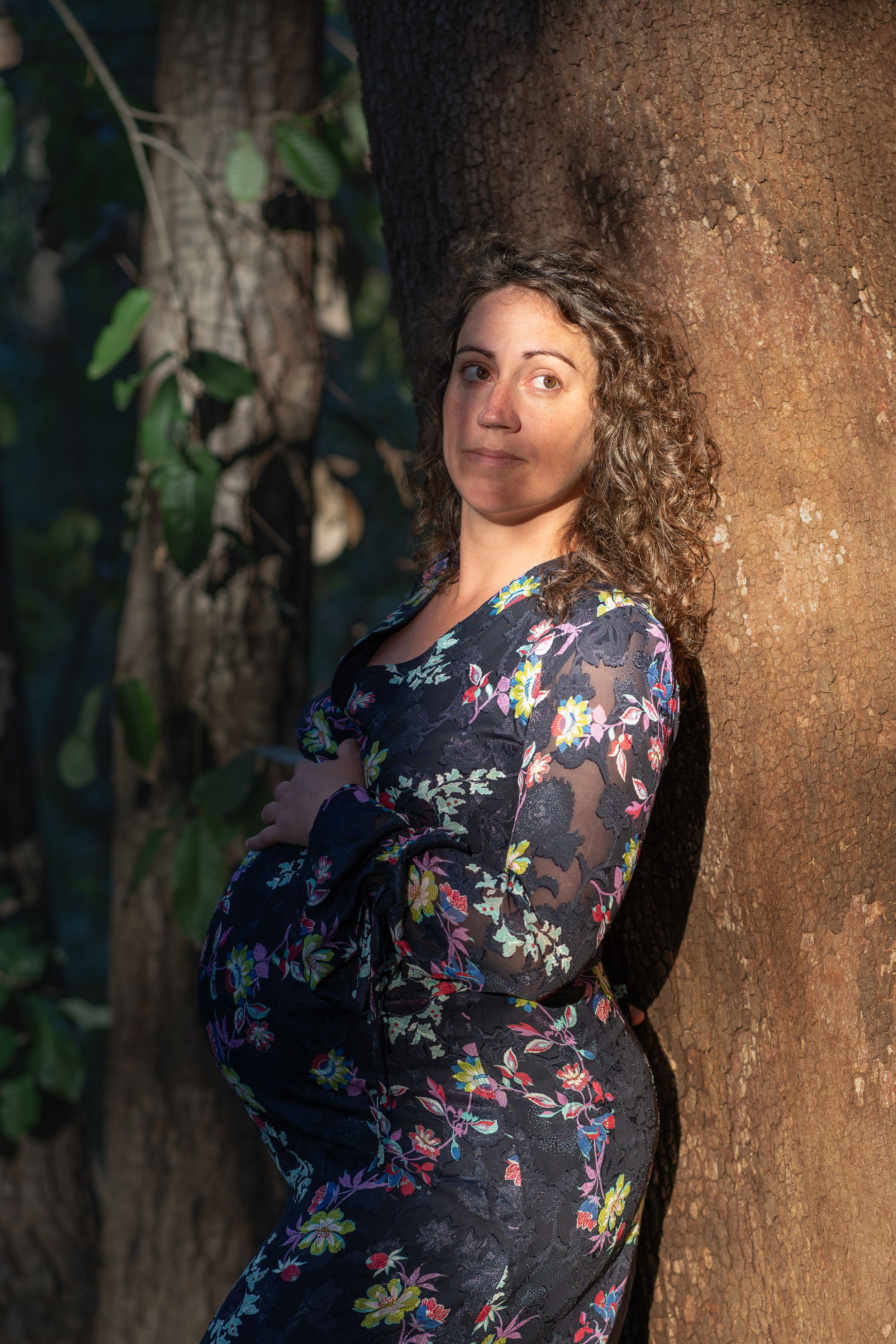 Maternity portrait of woman in floral dress leaning against tree trunk.