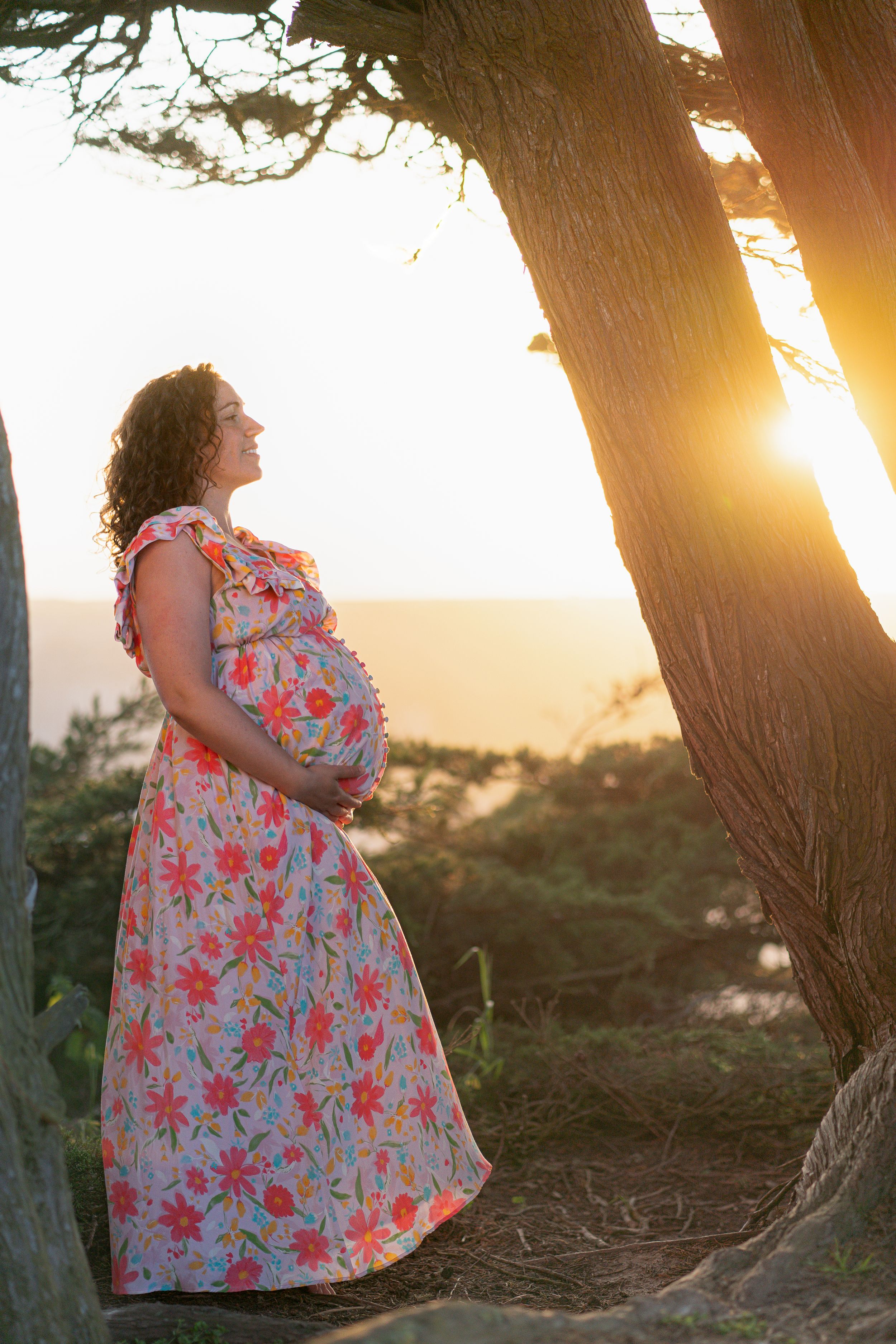 A pregnant woman in a floral dress stands beside a tree at sunset.
