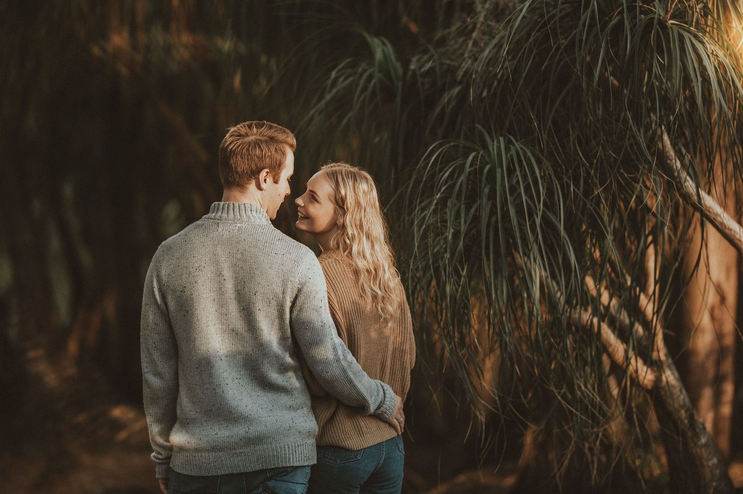 A couple in casual attire standing close together among plants during golden hour.