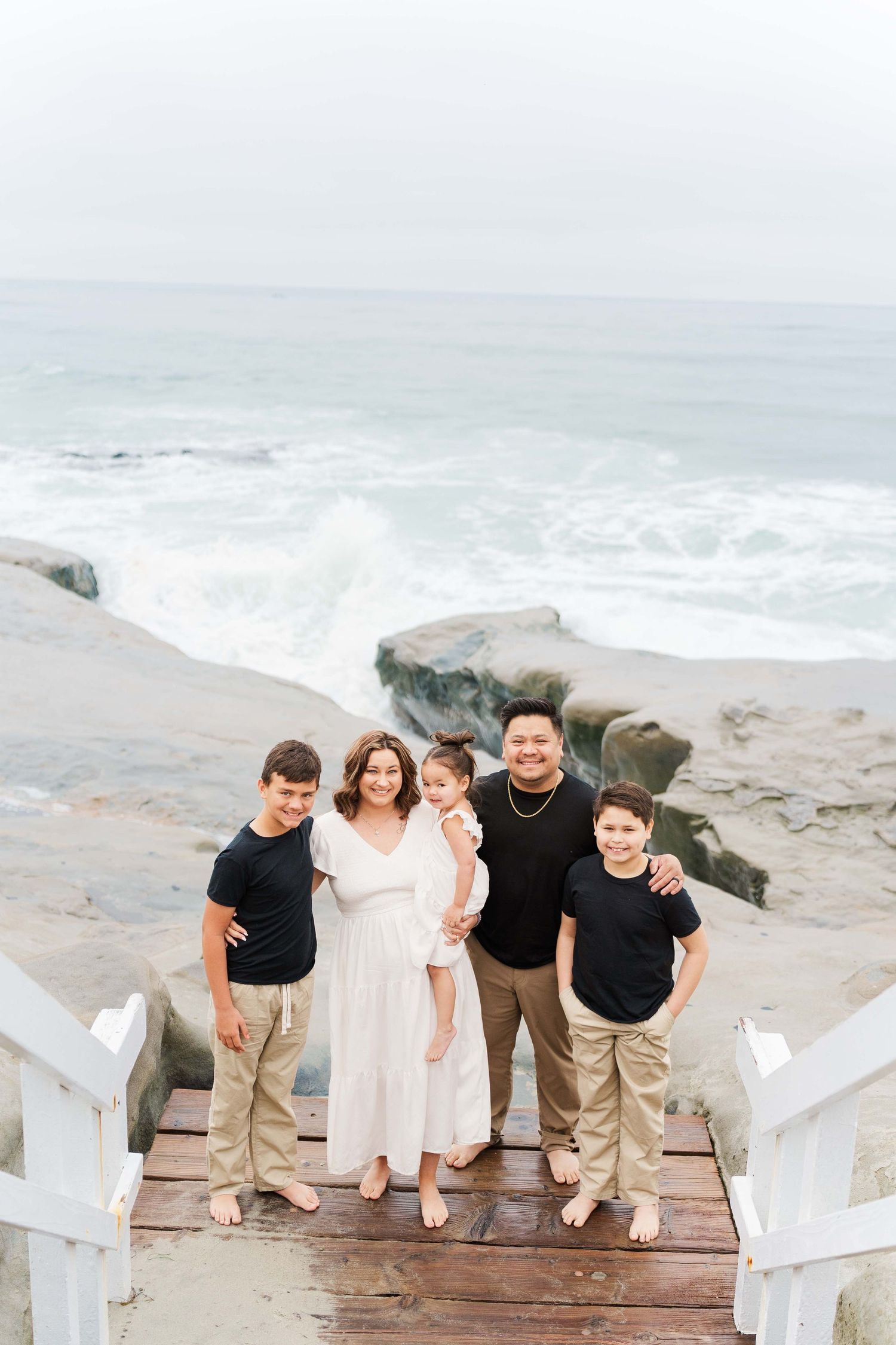 Family stands together on wooden stairs leading to rocky ocean shore on overcast day.