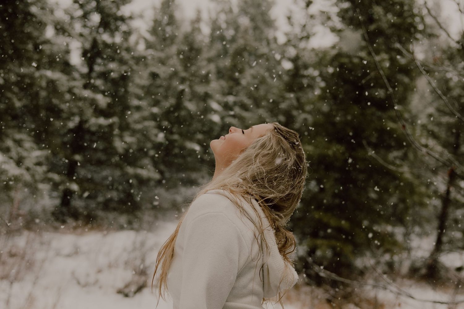 A person in a white sweater stands in a snowy forest looking up at falling snow during winter.