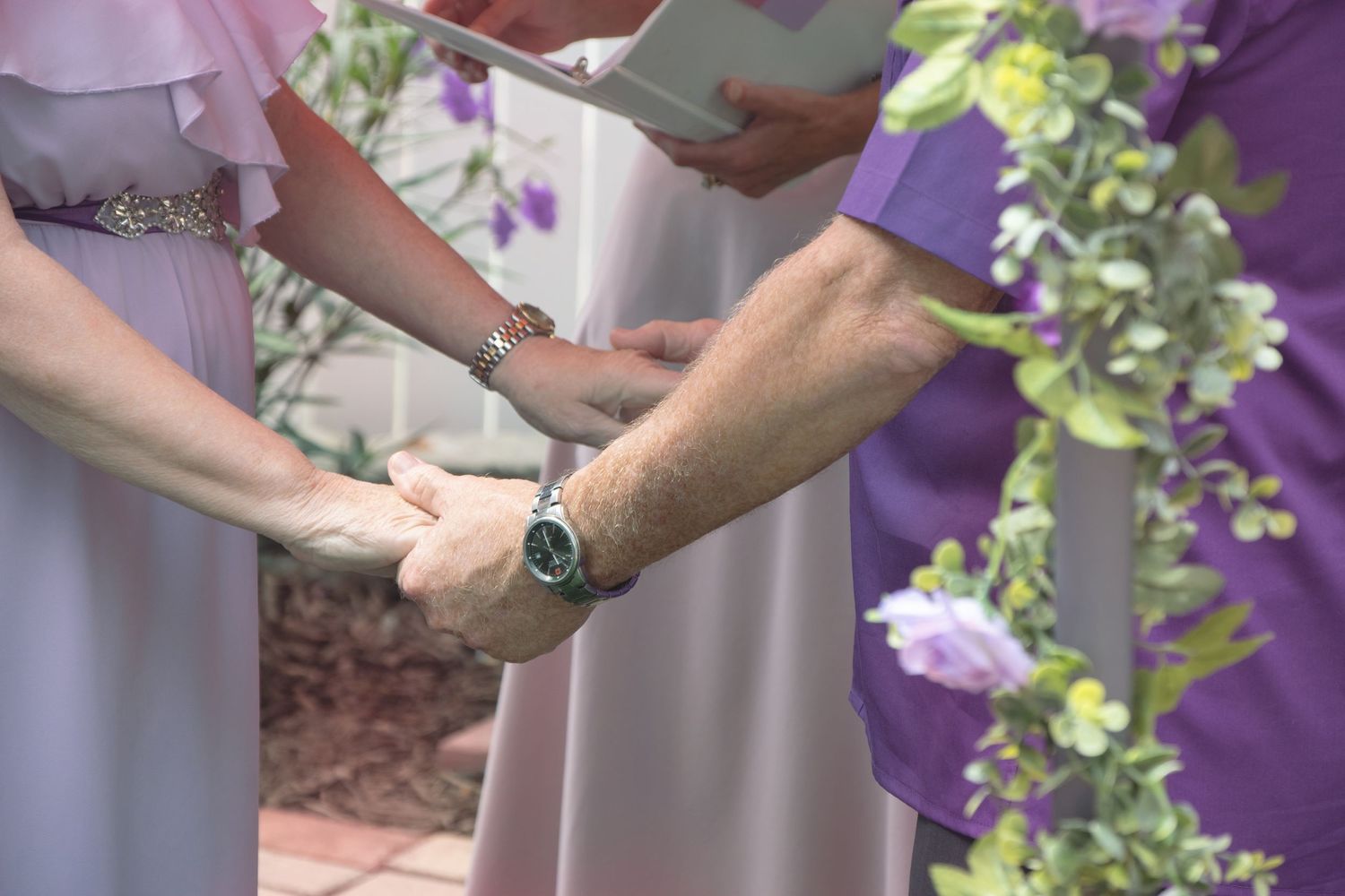 Hands joined together during an outdoor wedding ceremony with purple flowers in the background.