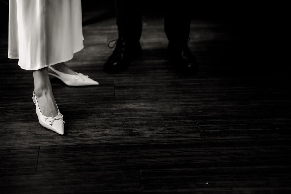 Black and white photograph showing elegant white shoes and dress hem against a dark wooden floor.