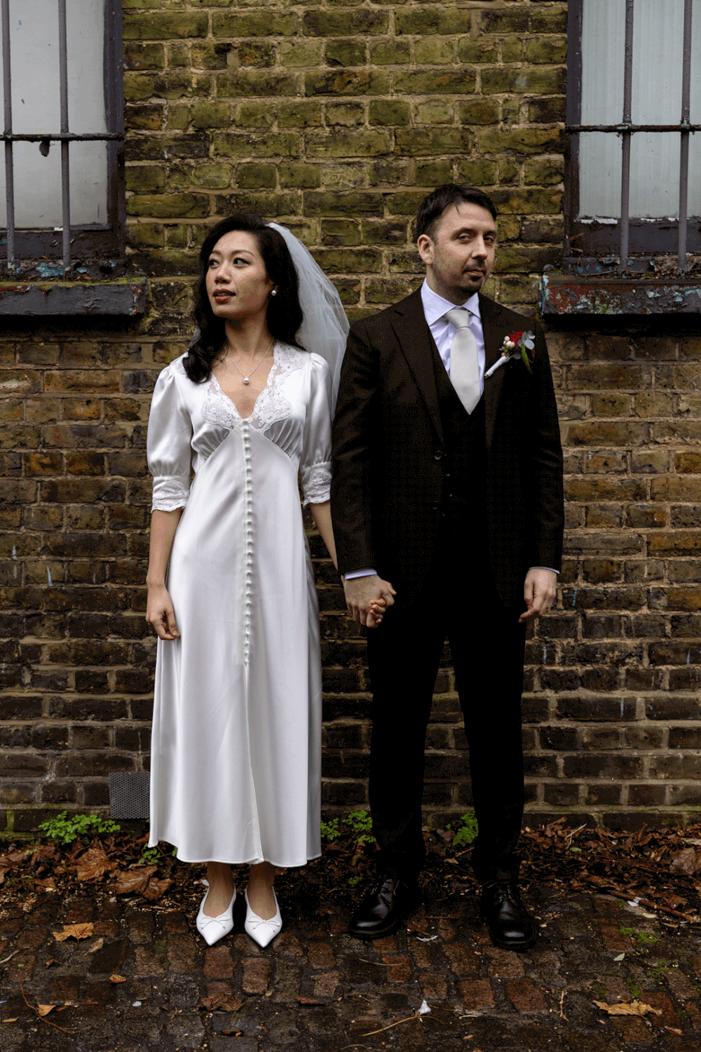 Bride and groom stand against rustic brick wall holding hands on their wedding day.