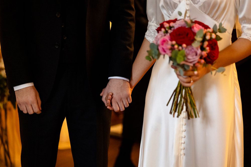 A bride holding a vibrant red and pink bouquet stands beside her groom, showing their wedding rings.
