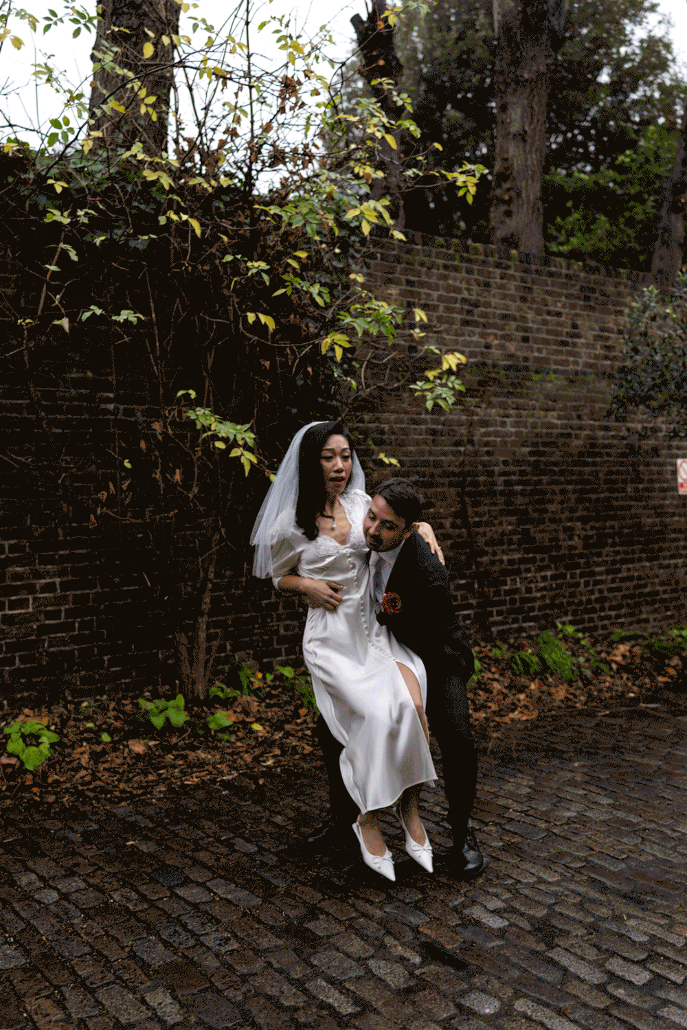 A couple embraces on cobblestone street against historic brick wall as sunlight filters through overhead trees.