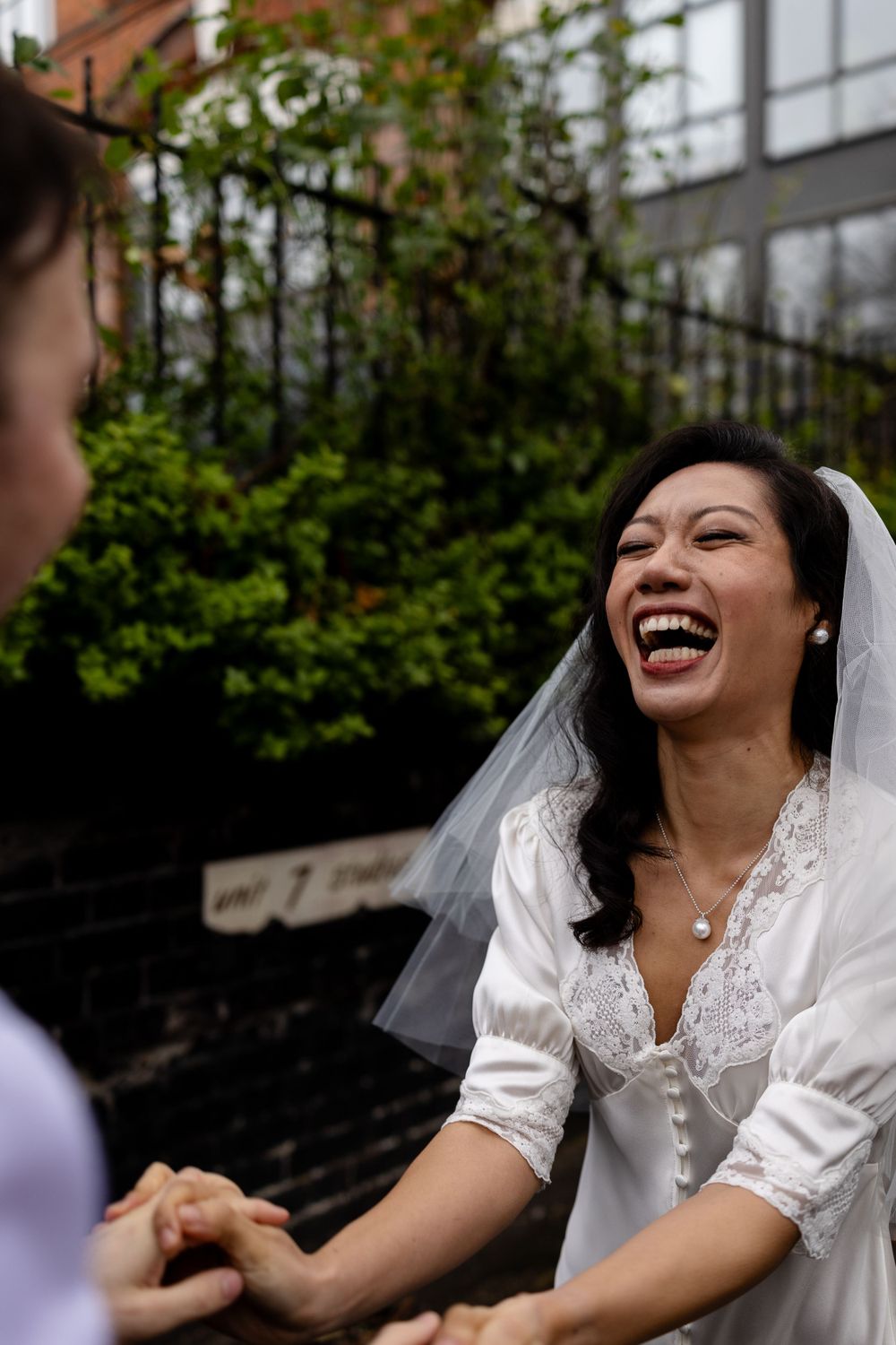 A joyful bride in a white wedding dress laughs heartily while holding someone's hands in an outdoor garden setting.