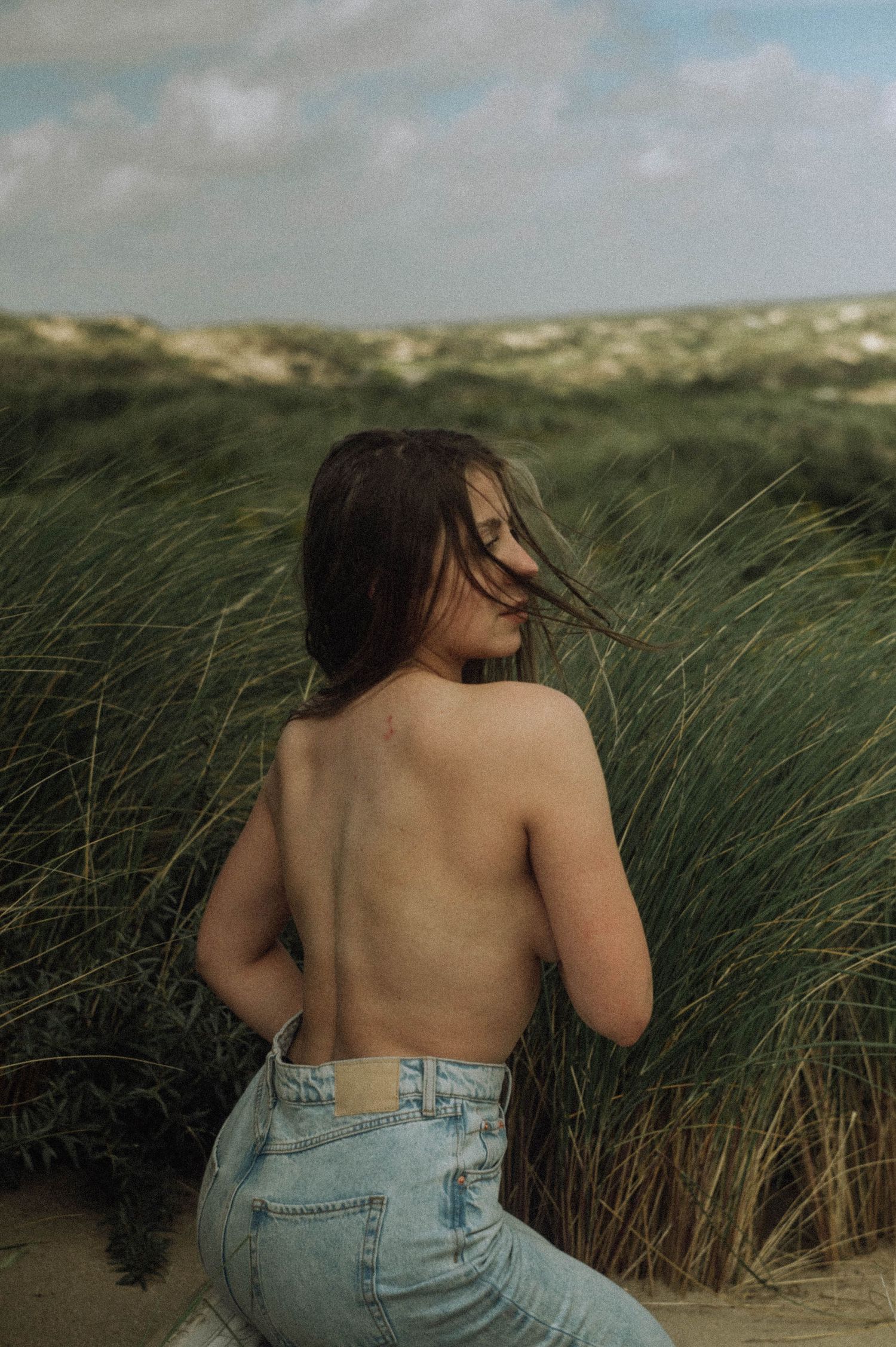 Boudoir photo in the dunes: a woman in jeans photographed topless from behind in nature.