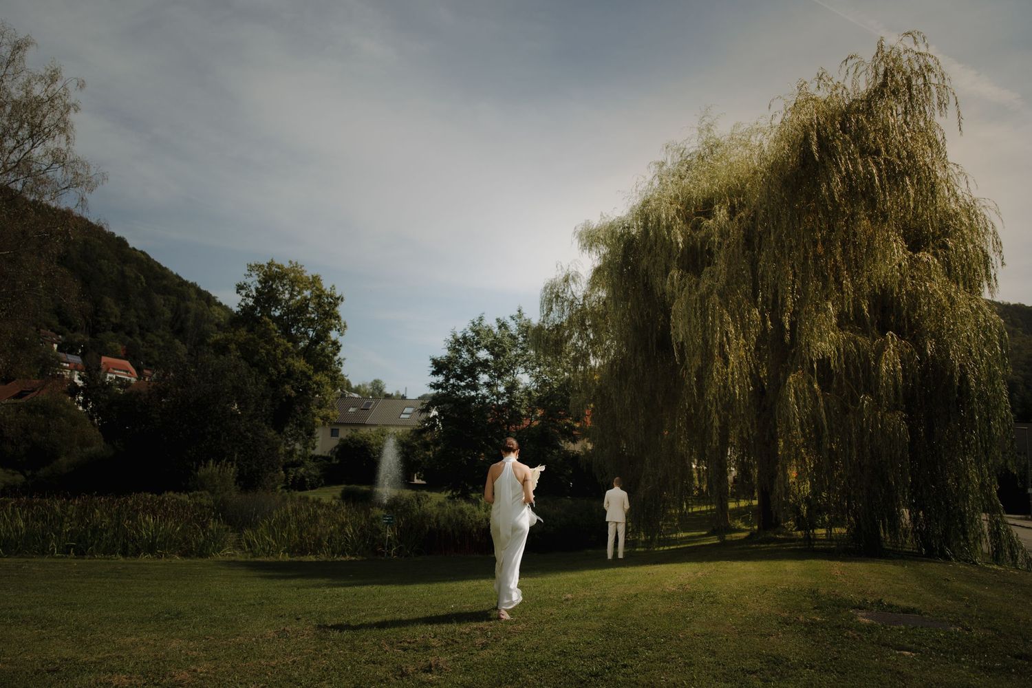 Two figures walking across a lush green lawn with weeping willow trees in the background.