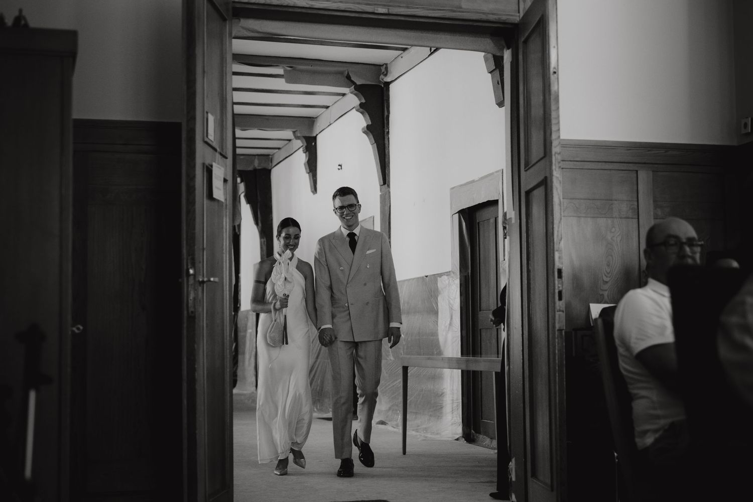 A couple walks through a hallway with white walls and wooden trim.