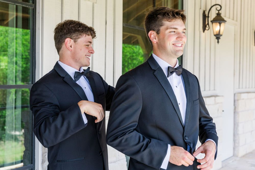 Two men in tuxedos and bowties prepare for a formal event on a covered porch.