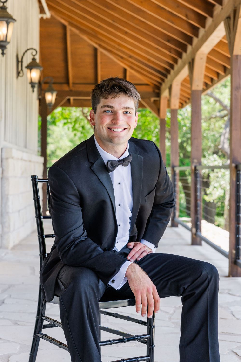 Groom in black tuxedo sitting on dark wooden chair on covered porch with rustic lanterns.