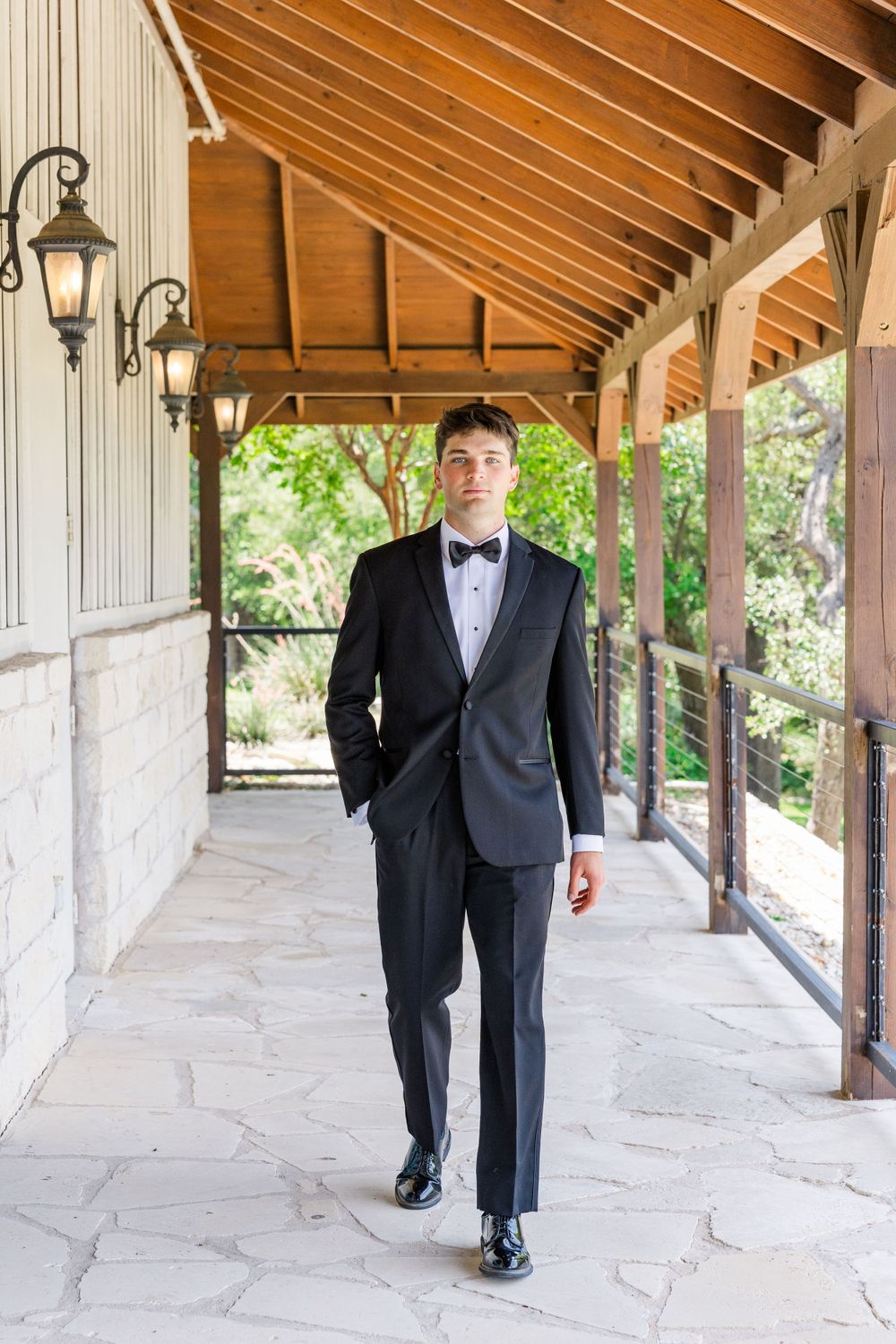 A person in a formal black tuxedo and bow tie walks along a covered stone porch with rustic wooden beams and lanterns.