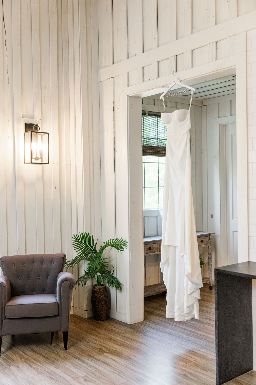 A white wedding dress hangs in a doorway of a modern room with cream paneled walls, gray armchair and hardwood floors.
