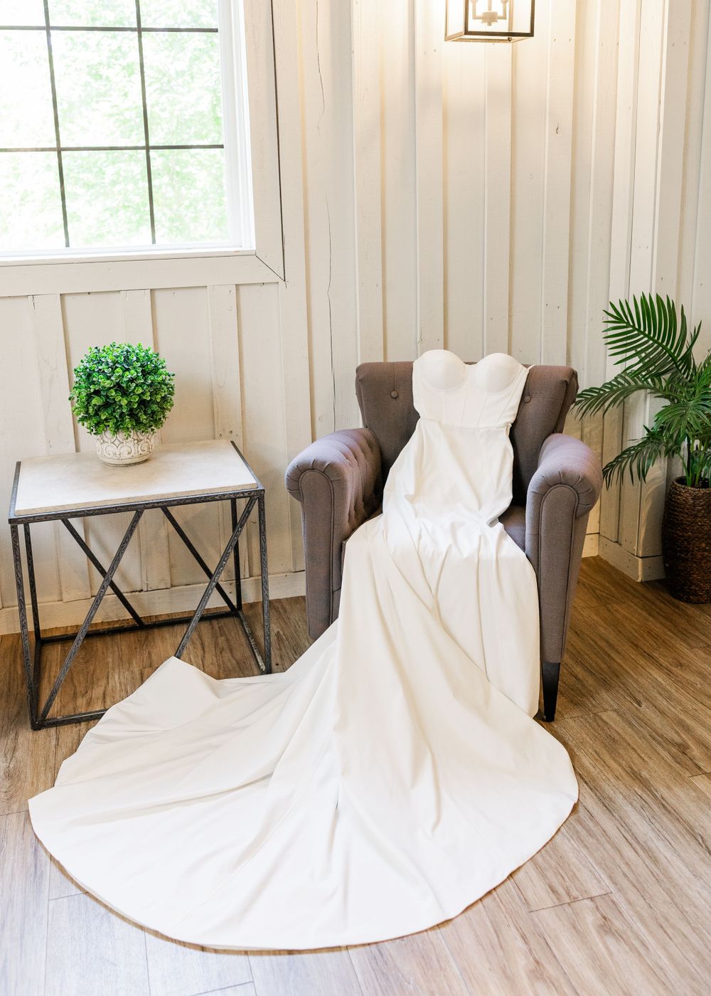 Elegant white wedding dress draped over brown armchair near window with side table and greenery accents.
