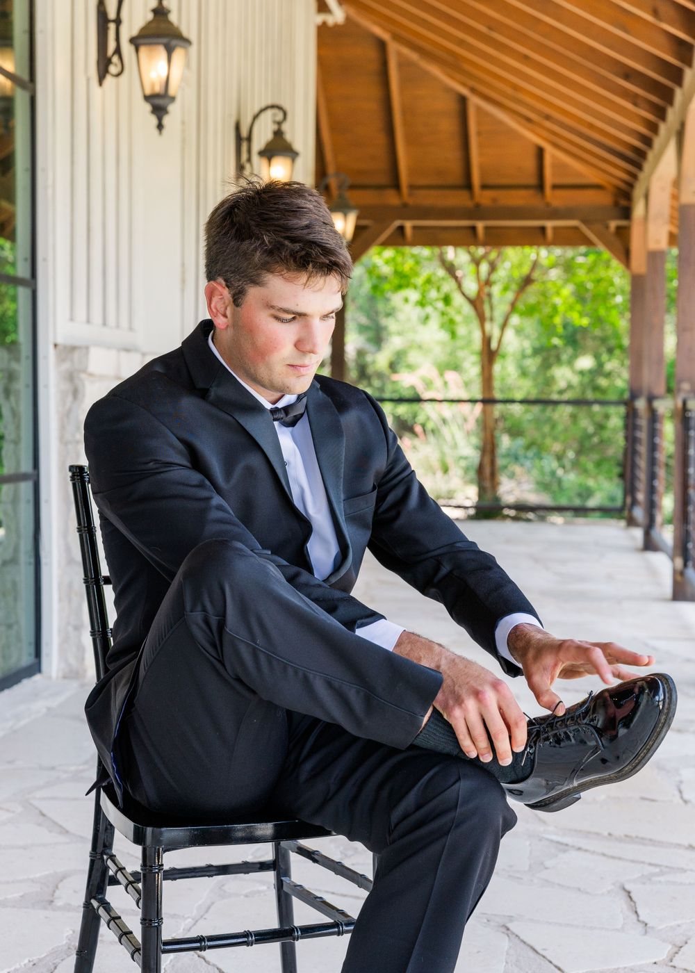 Person in formal black suit adjusts their shoe while sitting in a wooden rocking chair on a porch.