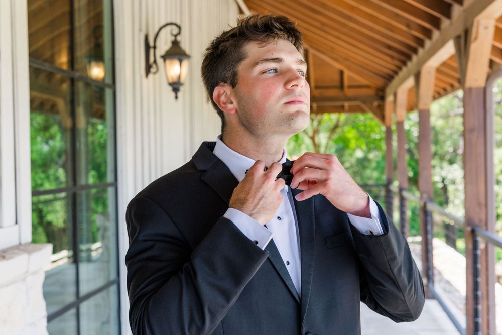 A man in formal attire adjusts his bowtie on a covered porch with lanterns in the background.