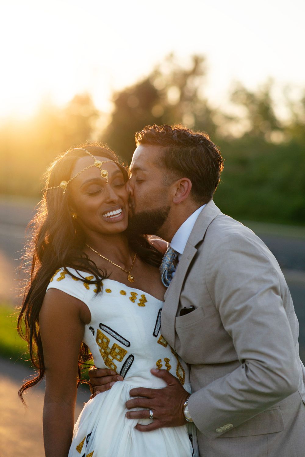 Golden hour wedding photo groom kissing bride on cheek with trees in the background. Bride in traditional Zuria. Sweet candid authentic warm