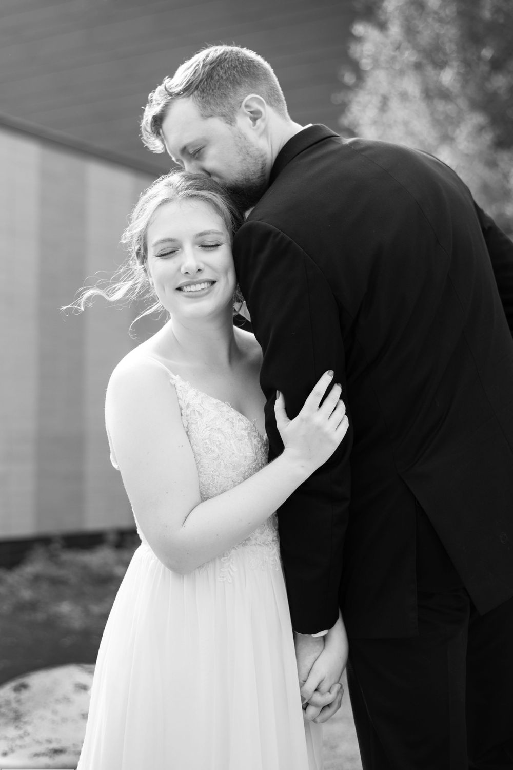 Classic black and white, timeless authentic sweet wedding photo. Groom kissing bride on top of head, bride hand on groom's arm. Romantic