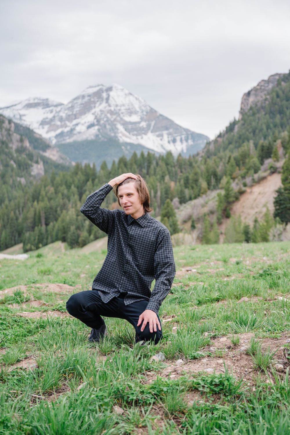 Person in dark sweater crouches on grassy hillside with snowy mountain peaks in distance.