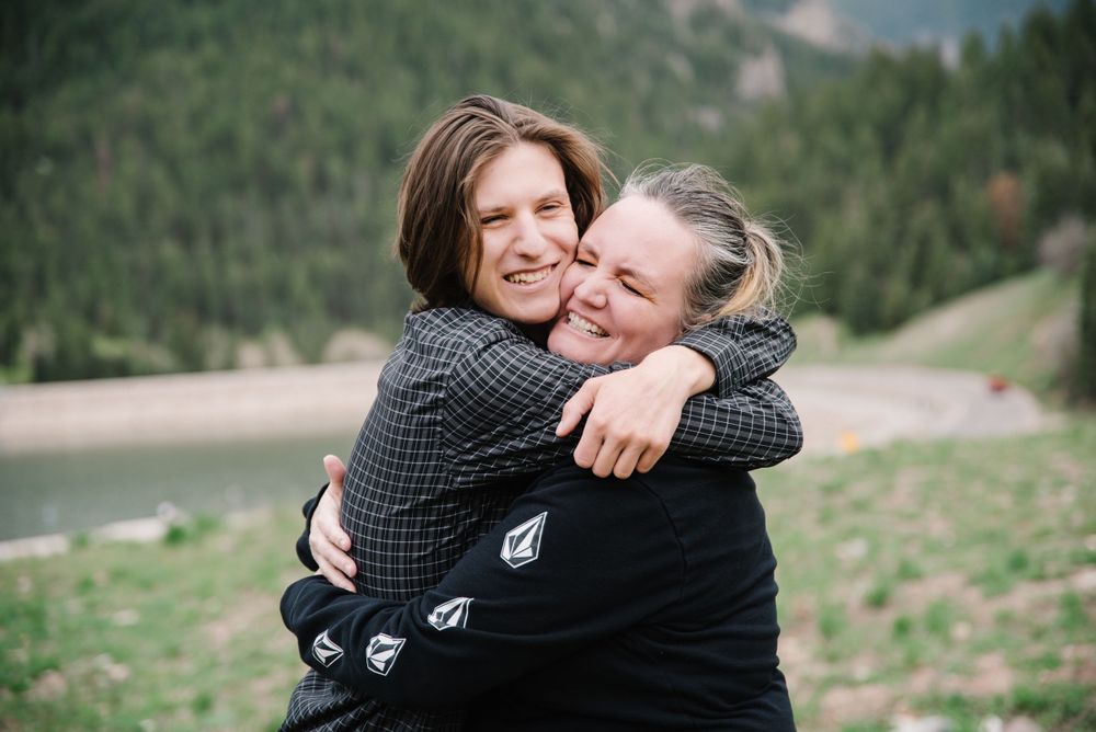 Two people share a joyful embrace outdoors with mountain scenery behind them.