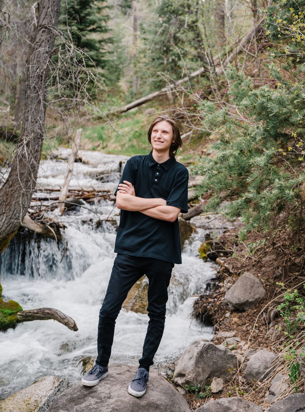 Person stands on rocky outcrop next to rushing mountain stream in forest setting.