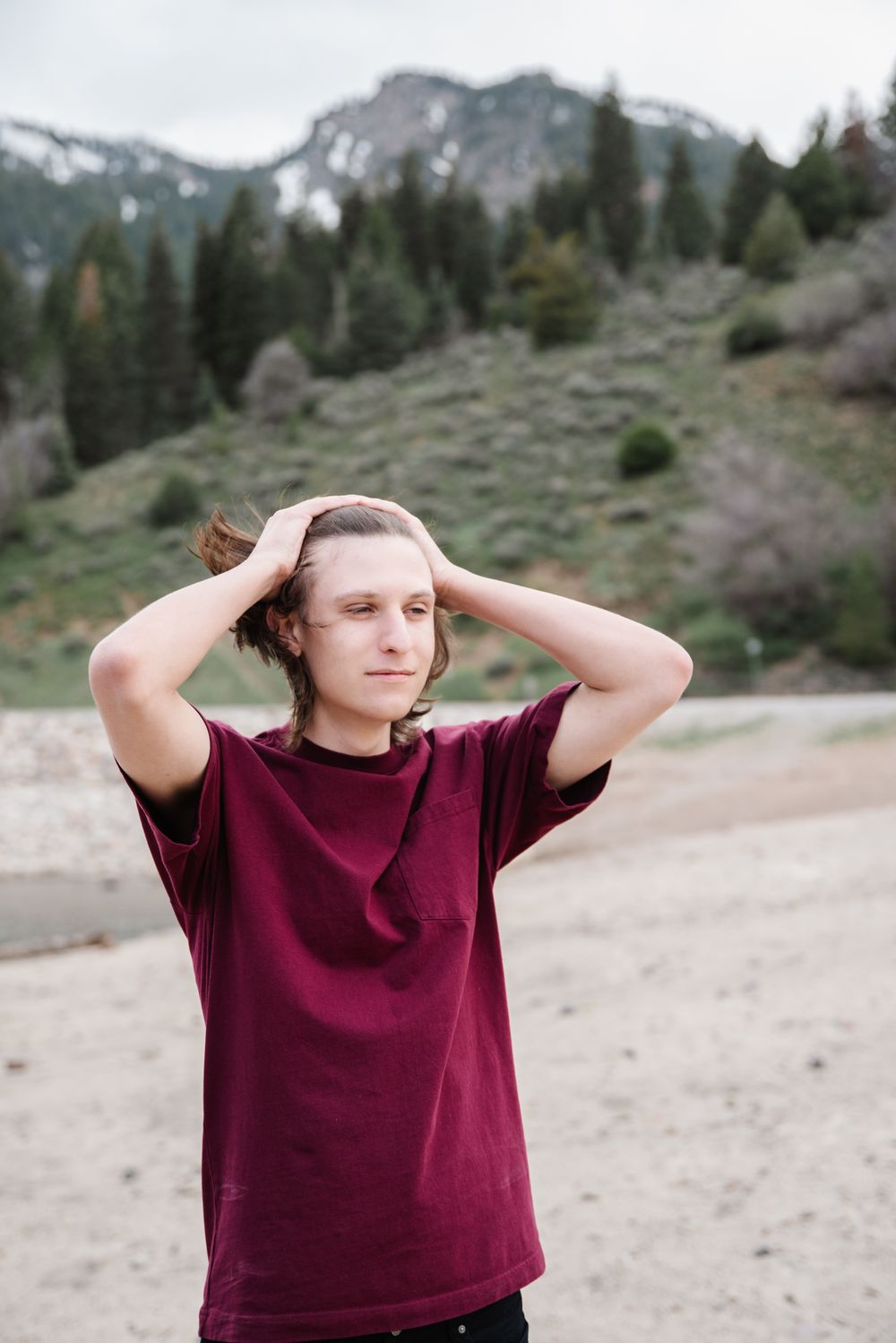 Person in burgundy shirt poses with hands behind head against mountain backdrop.