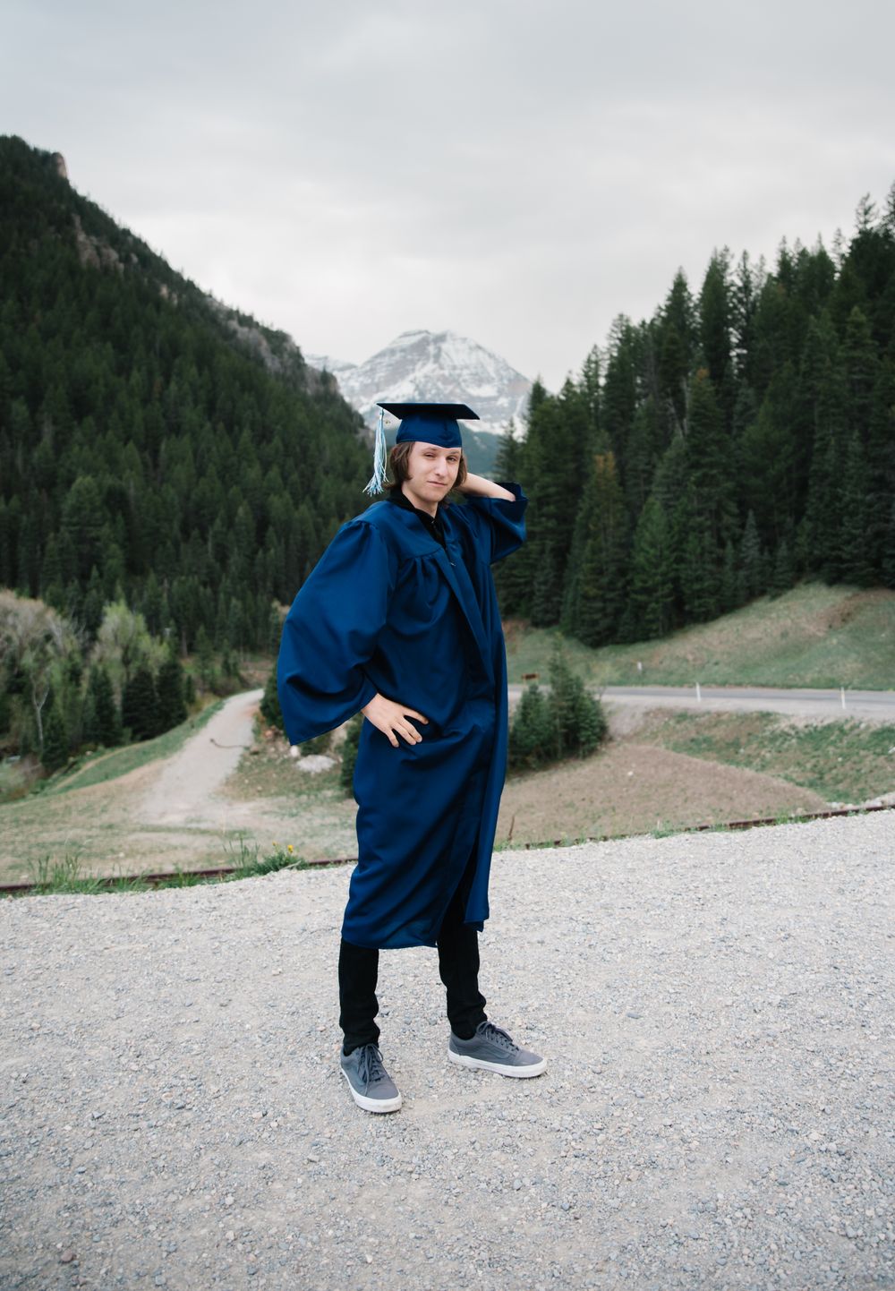 Graduate strikes playful pose in blue graduation gown with mountain valley in background.