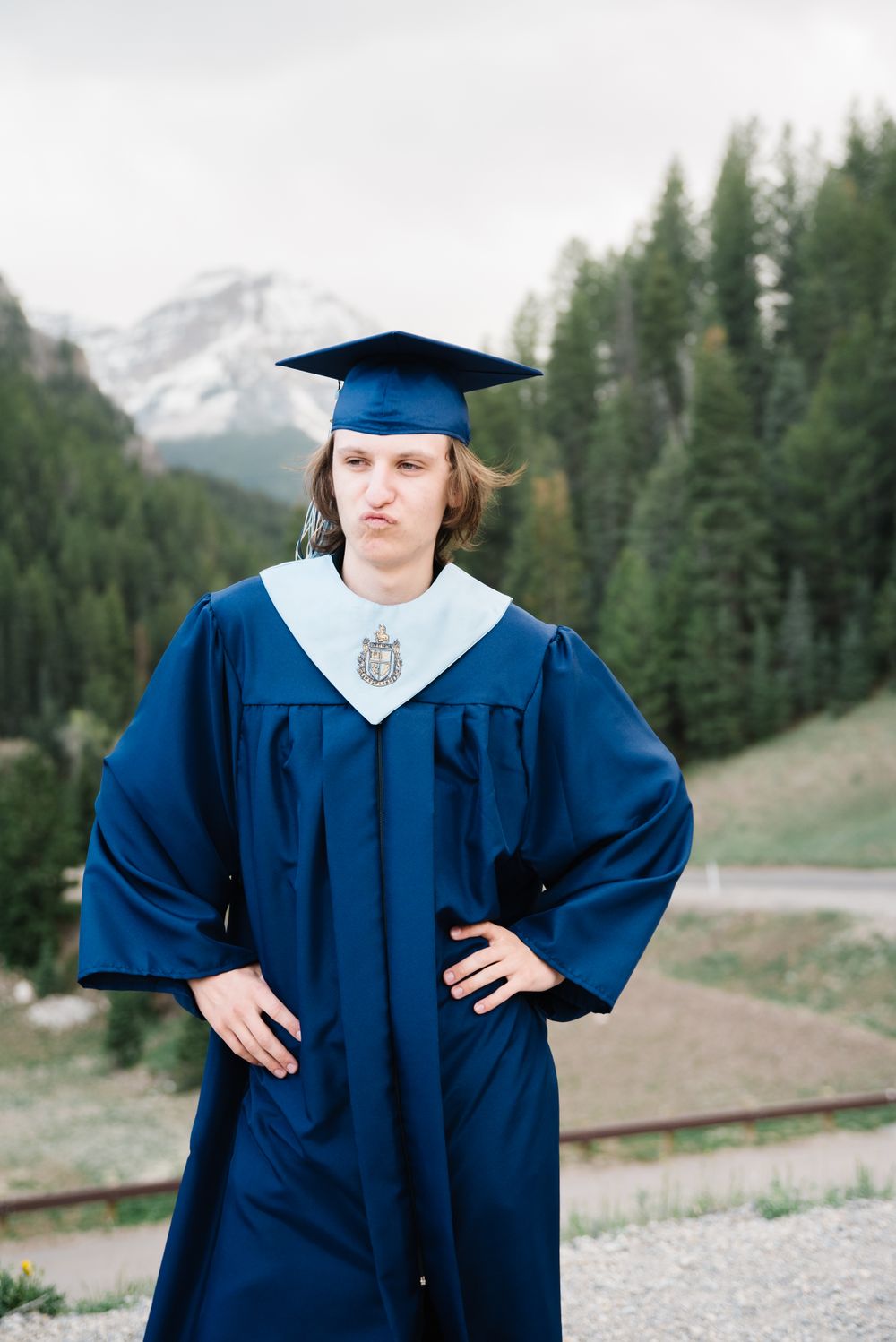 Graduate in blue cap and gown poses with snow-capped mountains and evergreen forest in background.