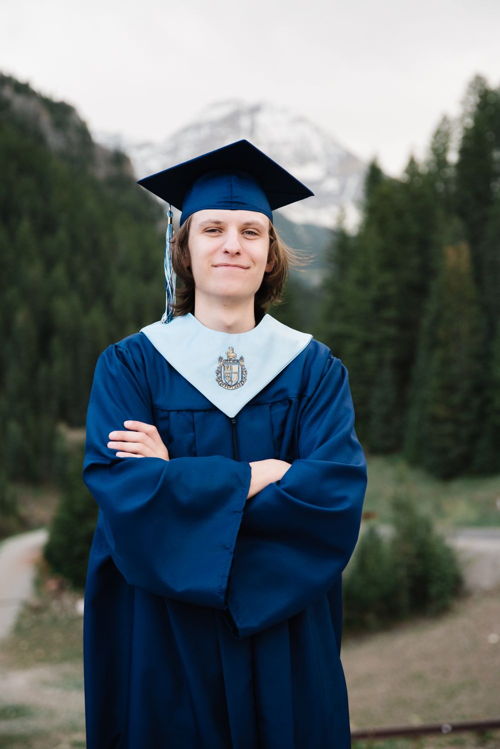 Graduate stands confidently in blue graduation attire against mountainous landscape.