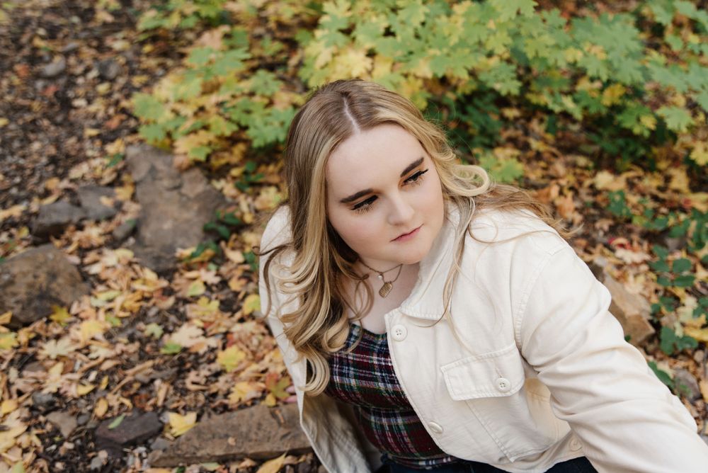 Person in white jacket and plaid dress sits among fallen autumn leaves.