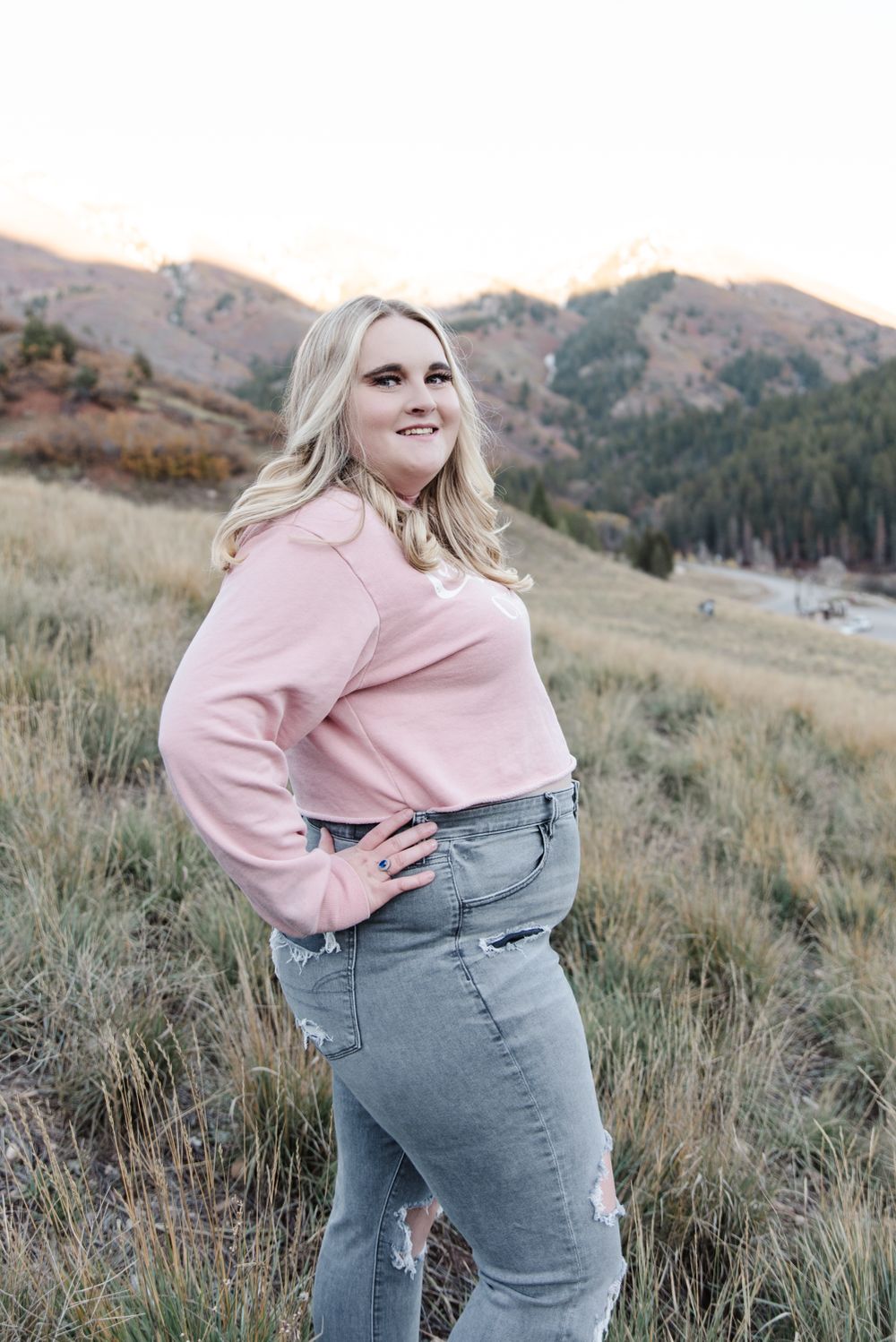 Person in pink sweater and jeans poses against mountainous autumn landscape.