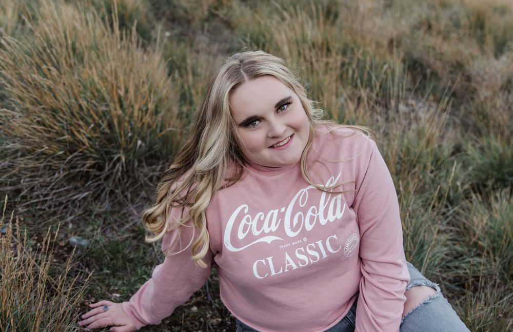 Woman in pink Coca-Cola Classic sweatshirt poses outdoors in natural grassland setting.